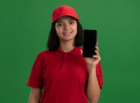 显示身穿红色制服 戴着帽子的年轻送货员站在绿色的墙上 脸上露出微笑立场移动脸