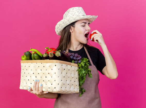 蔬菜穿着制服的漂亮园丁女孩戴着园艺帽 手里拿着菜篮子 咬着粉红色背景上孤立的西红柿花园女孩咬