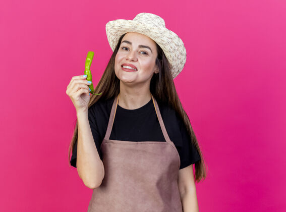 帽子微笑美丽的园丁女孩穿着制服 戴着园艺帽 手里拿着一个粉红色背景上孤立的破胡椒穿持有胡椒