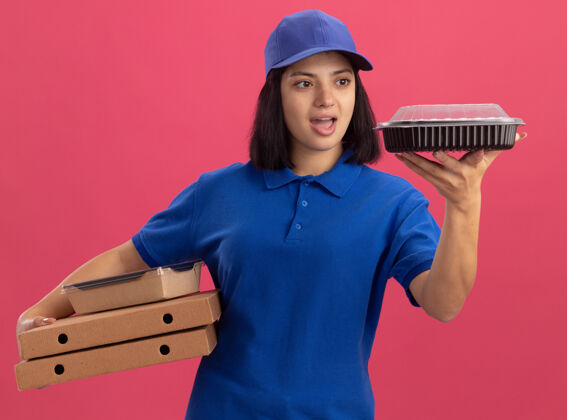 食品身穿蓝色制服 头戴鸭舌帽的年轻送货女孩拿着比萨饼盒和食品包 站在粉红色的墙上惊讶地看着包装年轻包装盒子