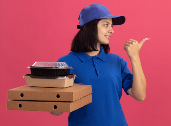食物身穿蓝色制服 头戴鸭舌帽 手持比萨饼盒和食品包的年轻送货女孩微笑着用拇指指着站在粉色墙上的一边包装拇指壁板