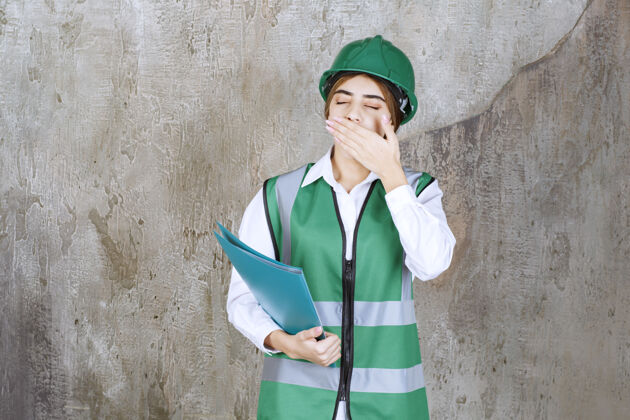 混凝土身穿绿色制服 头戴安全帽的女工程师手里拿着一个绿色项目文件夹 看上去又累又困建筑工程师女人