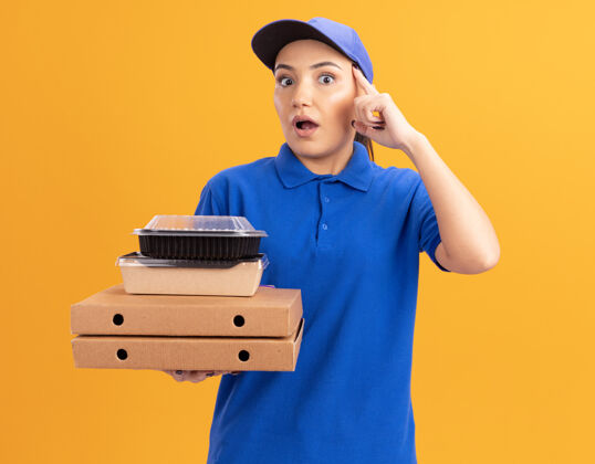 手指身穿蓝色制服 头戴鸭舌帽 拿着比萨饼盒和食品包的年轻送货员站在橙色的墙上 用食指指着自己的太阳穴 神情困惑盒子女人指着