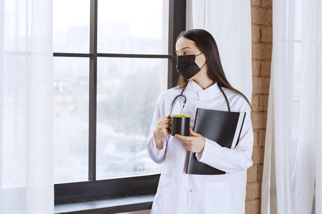 从业者医生拿着听诊器和黑色面罩 手里拿着一杯黑色饮料和一个黑色文件夹 从窗户往外看手术姿势水合
