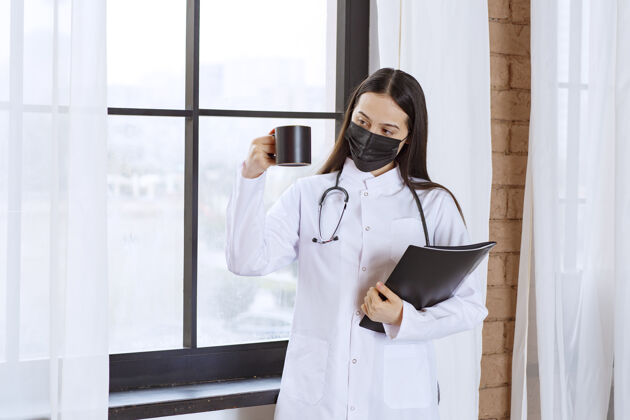 女性医生拿着听诊器和黑色面罩 手里拿着一杯黑色饮料和一个黑色文件夹放松报告治疗
