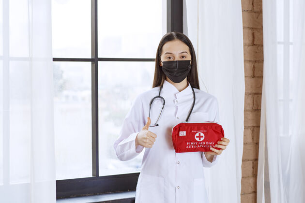 健康医生带着听诊器和黑色口罩 手里拿着一个红色的急救箱 竖起大拇指诊所从业者品牌