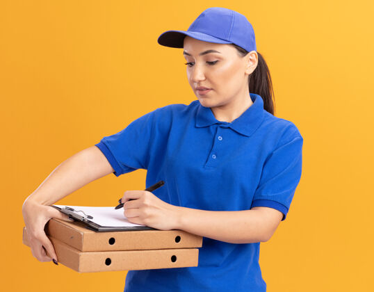 站着身穿蓝色制服 戴着帽子的年轻送货员 手里拿着比萨饼盒 脸上带着严肃的字迹站在橙色的墙上帽子女人披萨