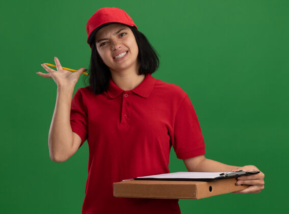 恼火身穿红色制服 头戴鸭舌帽 拿着比萨饼盒 拿着铅笔的年轻送货女孩站在绿色的墙上 脸上露出恼怒的表情披萨女孩年轻
