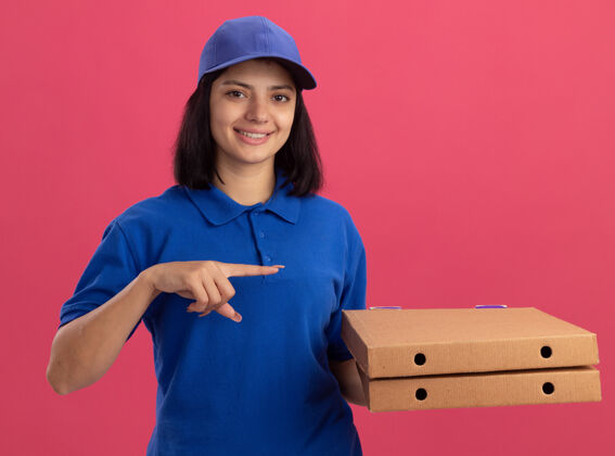 女孩身穿蓝色制服 头戴鸭舌帽 拿着披萨盒的年轻送货女孩 食指指着一旁微笑着站在粉色的墙上披萨送货手指