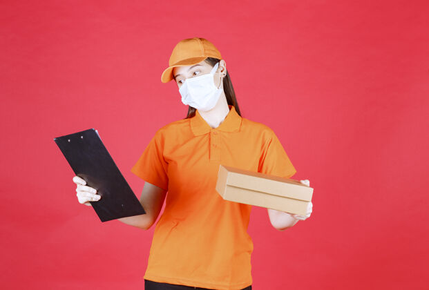 货物女信使身穿橙色制服 戴着口罩 手里拿着一个纸板箱 念着姓名和地址剪贴板制服检查单