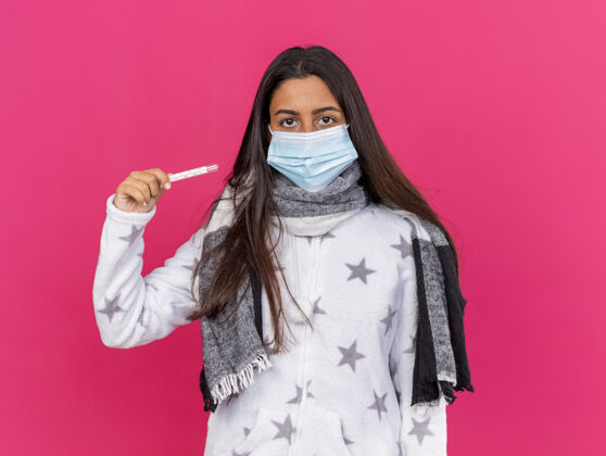 年轻年轻生病的女孩戴着医用面罩 戴着围巾 拿着隔离在粉色背景上的温度计 看着摄像机疾病穿相机