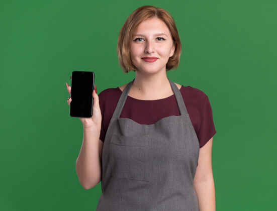 女人年轻漂亮的女理发师围裙展示智能手机微笑自信站在绿色的墙壁理发师微笑自信
