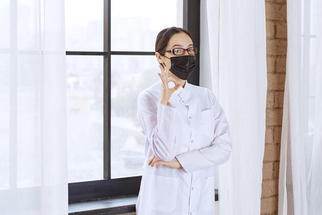 医学戴着黑面具和眼镜的医生站在窗边 展示着享受的标志模特成功病毒
