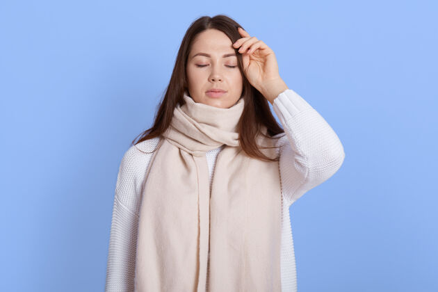 温度不健康妇女的画像裹着温暖的白围巾 摸着她的头 头痛 发烧和流感症状 眼睛紧闭 隔离在淡紫色的墙上女性病毒室内