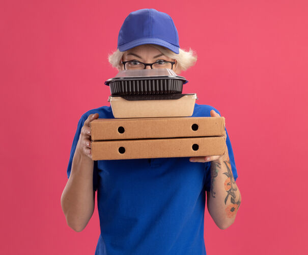 脸身穿蓝色制服 戴着眼镜 拿着比萨饼盒和食品包的年轻送货员 脸上带着严肃的表情 站在粉红色的墙上食物眼镜披萨