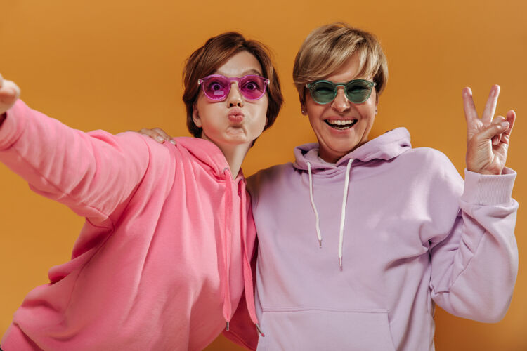 女士有趣的两位女士 现代短发 戴着炫酷的太阳镜 粉色和淡紫色的连帽衫 在橙色的背景下自拍 玩得很开心成熟年龄年轻