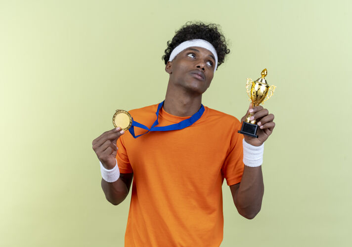 壁板看着一边思考的年轻的非裔美国运动男子戴着头带和腕带 拿着奖牌杯孤立的绿色背景头带运动腕带