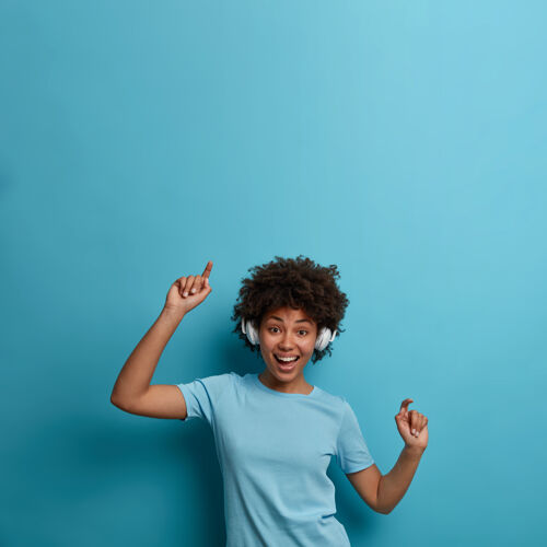 享受积极快乐的美国黑人年轻女子喜欢音乐的调子 戴着无线耳机 随着歌曲的节奏移动 有快乐的心情 隔离在蓝色的墙上 复制上面的空间休息 生活方式非洲乐观舞蹈