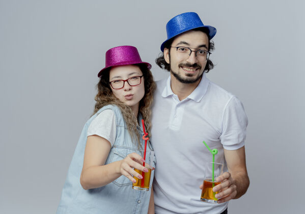 年轻高兴的年轻夫妇戴着粉红色和蓝色的帽子 拿着一杯果汁隔离在白色背景上情侣果汁戴着