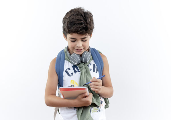 衣服高兴的小男孩戴着背包和耳机在笔记本上写东西隔离在白色的背景小耳机写字