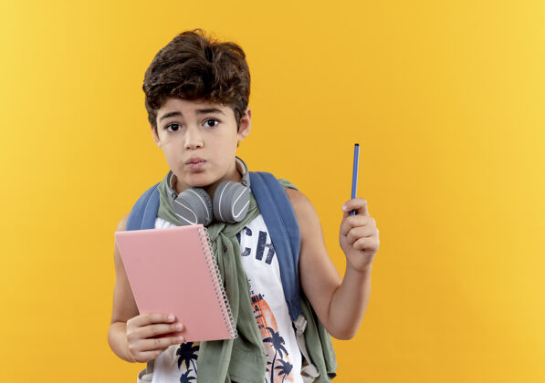书包迷茫的小男孩戴着背包和耳机拿着笔记本 黄色背景上的笔是孤立的小耳机男孩