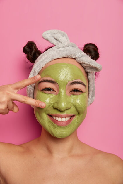 泥在美丽的过程中 自然亚洲女人的近景拍摄有乐趣 在眼睛上做和平手势 应用绿色净化面膜 清洁皮肤 在粉红色墙壁上赤身裸体站立应用微笑请