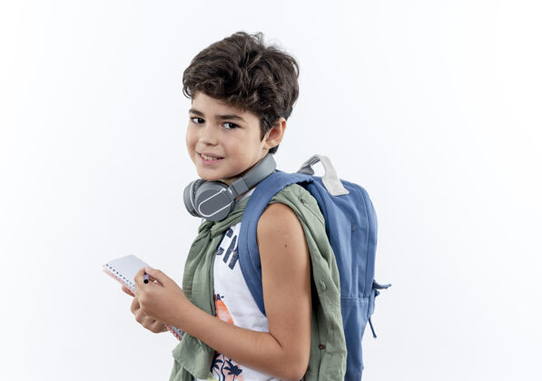 包高兴的小男生背着书包戴着耳机拿着笔记本和笔隔离在白色背景上小的背笔记本