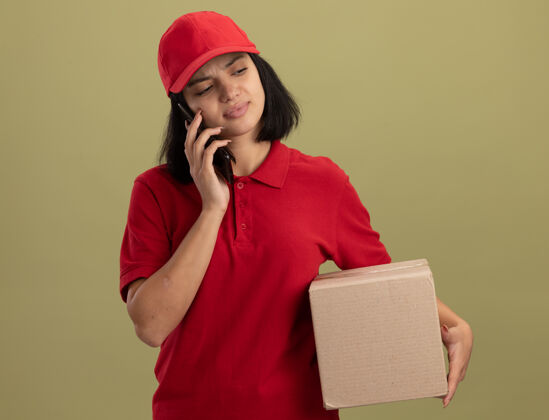 困惑身穿红色制服 头戴鸭舌帽的年轻送货员拿着纸板箱讲着手机 站在灯火通明的墙上感到困惑和不快手机拿着灯