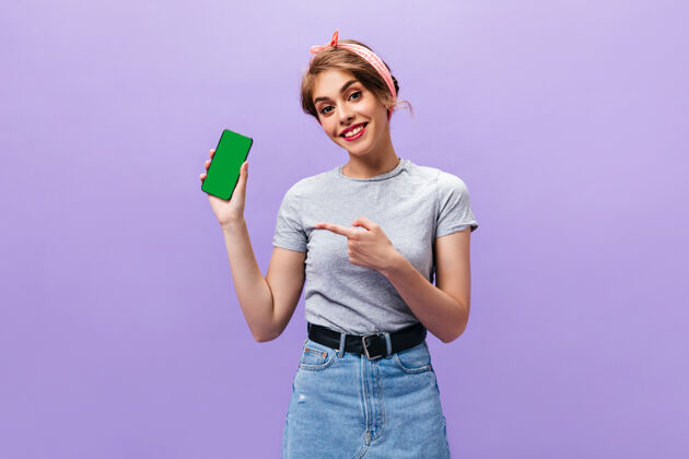 空间身穿灰色衬衫的年轻女子在紫色背景上演示手机迷人的女孩在时尚的衣服和粉红色的头带微笑头发休闲姿势