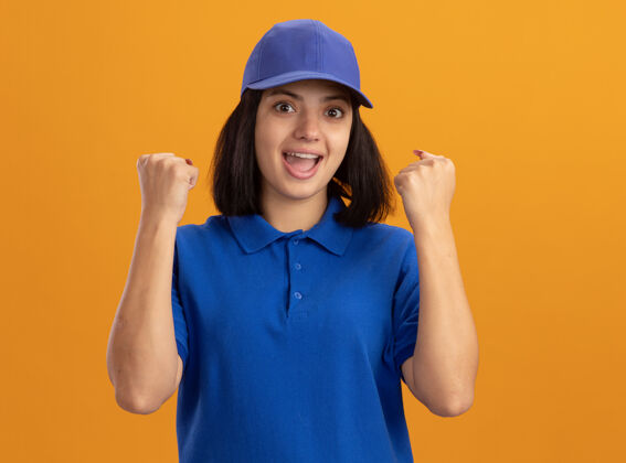 幸福身穿蓝色制服 戴着帽子 握紧拳头的年轻送货员站在橙色的墙上 快乐而兴奋年轻分娩拳头