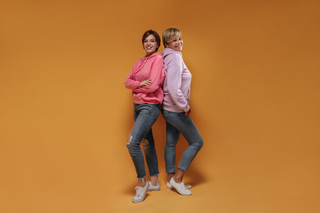 衬衫积极的两位短发女士 穿着时髦的粉色运动衫 时髦的牛仔裤和酷酷的运动鞋 面带可爱的笑容 在橙色的背景下看着镜头成人女士女士