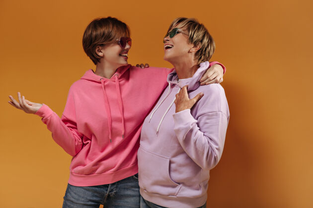 中等短发女人戴着太阳镜 穿着现代的淡紫色和粉色连帽衫 牛仔裤 在橙色孤立的背景下微笑着拥抱着休闲牛仔粉色