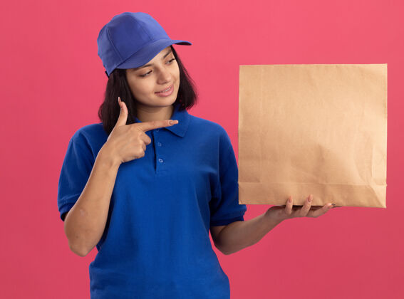 显示身穿蓝色制服 戴着帽子的年轻送货女孩站在粉红色的墙上 用食指指着纸包 自信地微笑着制服手指信心