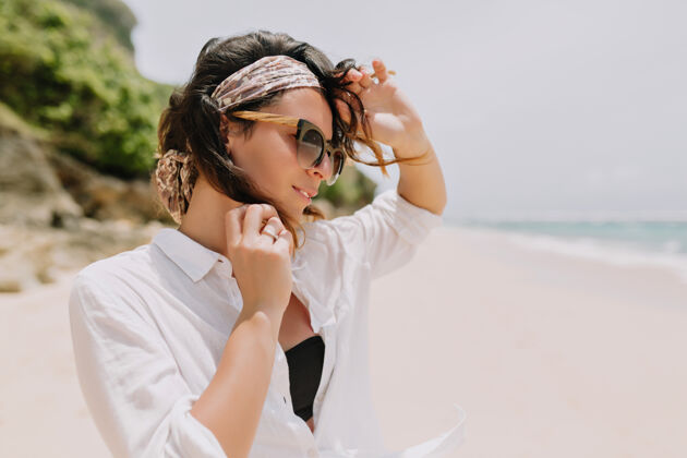 女性可爱可爱的女人 深色波浪发 穿着白色衬衫 戴着黑色太阳镜 在海边的白色沙滩上尽情享受 带着可爱的微笑沙滩海洋休闲