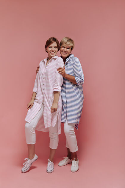 粉色背景快乐的两位女士 短发 条纹长衫 白色长裤 酷酷的运动鞋 在孤立的背景下微笑着拥抱着女性年轻老年人