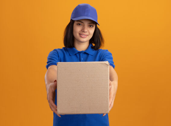 交货身穿蓝色制服 戴着帽子的年轻送货女孩站在橙色的墙上 脸上挂着微笑 手里拿着纸盒年轻微笑女孩