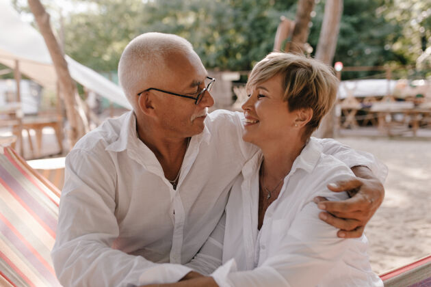 祖父母灰色头发的男人戴着眼镜 穿着长袖衬衫 拥抱着 看着海滩上穿着白色衬衫的短发微笑的妻子老年人退休海滩