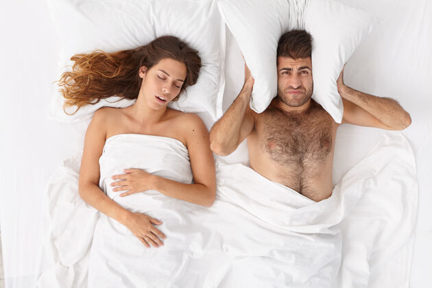 问题沮丧的男人用枕头捂住耳朵 因为妻子打鼾太大声而无法入睡 失眠 在卧室摆姿势疲倦的男人被女人打鼾激怒人 健康 睡眠障碍打扰婚礼愤怒
