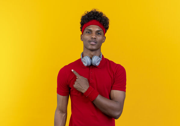 运动高兴的年轻黑人美国运动男子戴着头带和腕带与耳机点在一边隔离黄色背景与复制空间壁板点头带