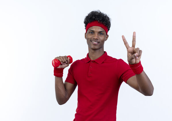 头带高兴的年轻黑人美国运动男子戴着头带和手环举着哑铃 并显示出和平的姿态隔离在白色背景上握运动穿
