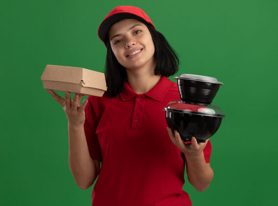站着身着红色制服 头戴鸭舌帽 手持食品包装 微笑友好地站在绿色墙壁上的快乐少女微笑女孩送货