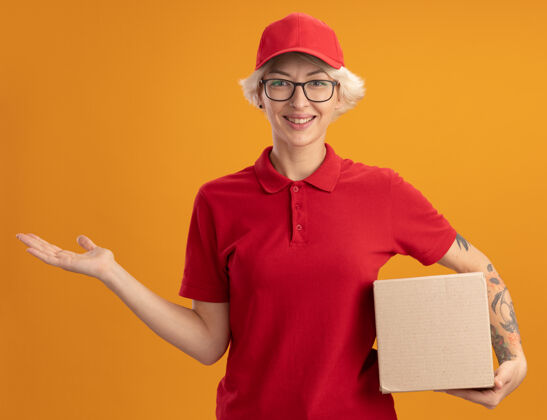 女人身着红色制服 戴着帽子 戴着眼镜 拿着纸板箱 微笑着展示复印空间的快乐年轻女快递员 她的手臂站在橙色的墙上帽子交货微笑