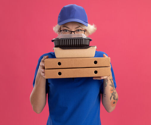 脸身穿蓝色制服 戴着眼镜 拿着比萨饼盒和食品包的年轻送货员 脸上带着严肃的表情 站在粉红色的墙上食物眼镜披萨