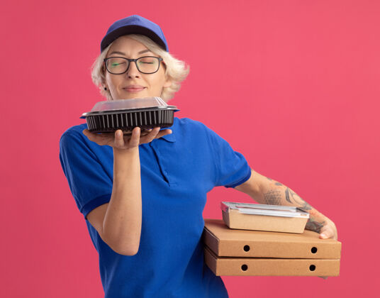 持有身穿蓝色制服 戴着眼镜 手持比萨饼盒和食品包装的年轻送货员在粉红色的墙上呼吸着令人愉快的香气披萨年轻眼镜