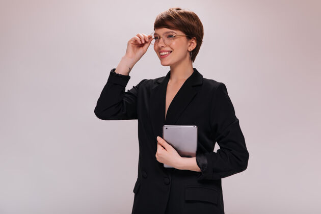 优雅戴眼镜的时髦女人微笑着拿着平板电脑穿着黑色西装的迷人女人在与世隔绝的背景下摆出好心情肖像时尚女性