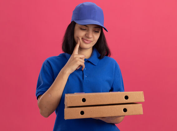 思考穿着蓝色制服 戴着帽子 拿着披萨盒的年轻送货女孩站在粉红色的墙上看着他们思考盒子年轻拿着