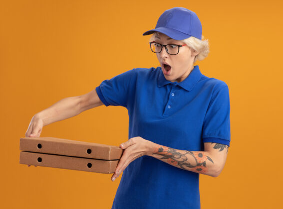 穿身穿蓝色制服 戴着眼镜 拿着比萨饼盒的年轻送货女在橙色的墙上惊讶地看着比萨饼盒女人制服拿着