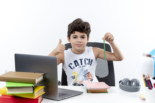 铅笔高兴的小男孩坐在书桌旁 手里拿着铅笔 他的大拇指孤立地竖立在白色的背景上拿着请工具