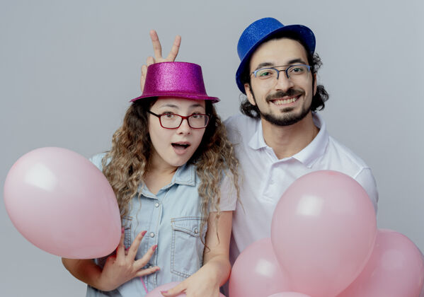 手势快乐的年轻夫妇戴着粉红色和蓝色的帽子做兔子耳朵的家伙手势女孩举行气球隔离在白色背景女孩男孩耳朵