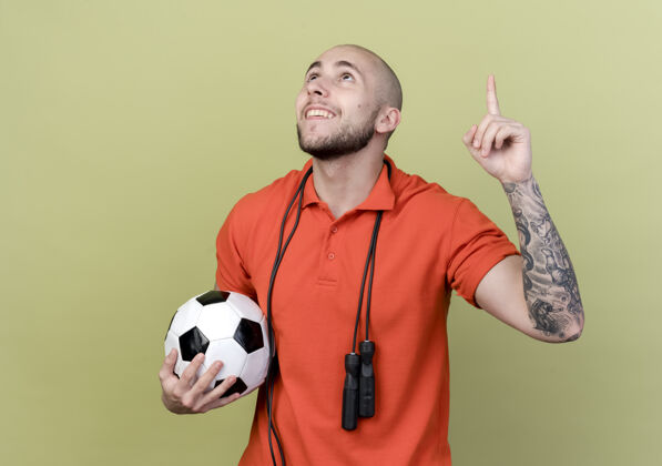 年轻仰望微笑的年轻运动型男子指着肩上举着跳绳的橄榄绿背景球运动微笑球